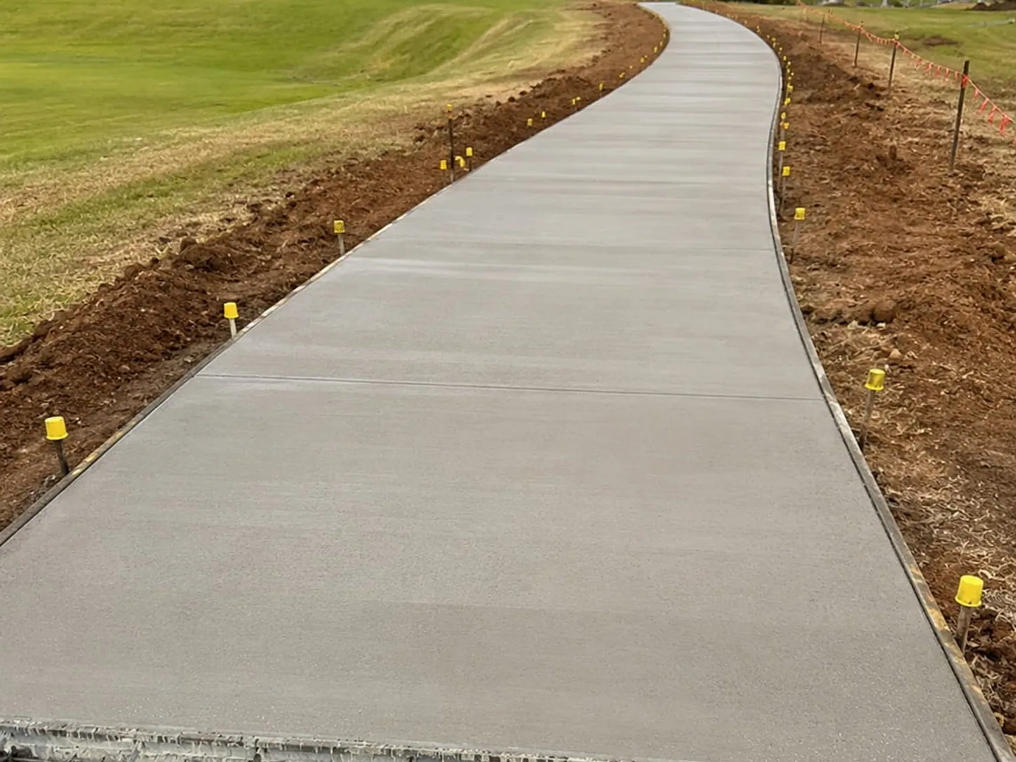 Wide concrete pathway curving across a lawn, freshly poured with the edge pegs still in place