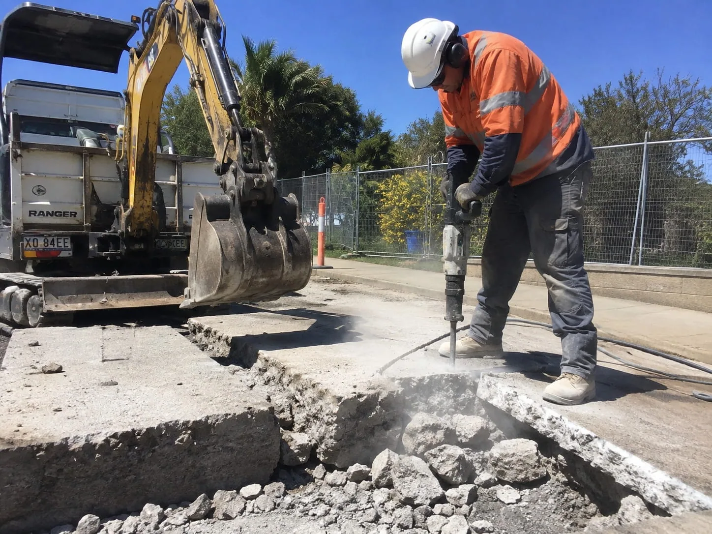 Worker breaking up an old concrete slab with a jackhammer while a mini excavator waits to clear the broken sections
